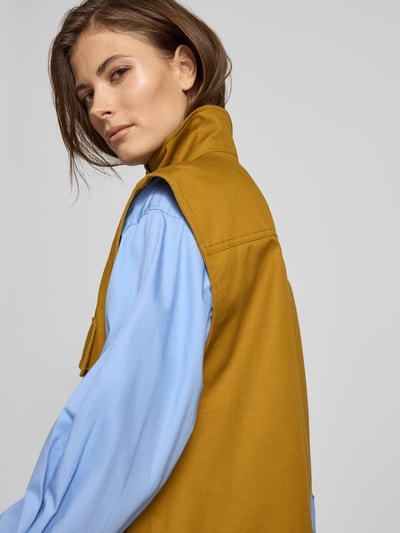 Modelka kobieca nosi Oversized Weste mit aufgesetzten Taschen - curated by Tinvcb od curated by Tinvcb, damska weste, krój relaxed fit, stójka, zapięcie na guziki, kieszenie z patką, długość pleców w rozmiarze 36: 67 cm, szerokość ramion w rozmiarze 36: 52 cm, szerokość w klatce piersiowej w rozmiarze 36: 59 cm, kolor musztardowy z jasnoniebieską koszulą, tekstura gładka, detaliczne wykończenie. Ujęcie z tyłu.