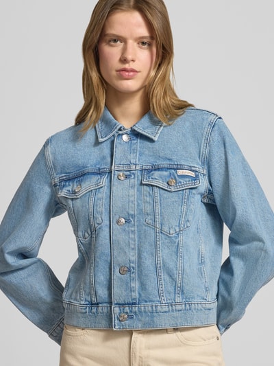 Vrouwenmodel draagt Regular Fit Jeansjacke met borstzakken model 'Panama' van Calvin Klein Jeans, elastische katoenmix, lichte denimkleur, knoopsluiting, logo-patch, opgestikte zakken, omgeslagen kraag, detailopname bovenlichaam.