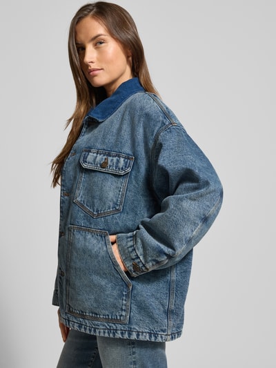 Frauenmodel trägt Kurtka jeansowa z bawełny o kroju relaxed fit, model 'JOYBIRD' od American Vintage, styl vintage, wpuszczane i kieszenie na piersi, wykładany kołnierz, listwa guzikowa, niebieska, faktura denim, długość tyłu 61 cm, szerokość ramion 56 cm, szerokość klatki piersiowej 68 cm, długość rękawów 55 cm, Ganzkörperansicht von vorne.