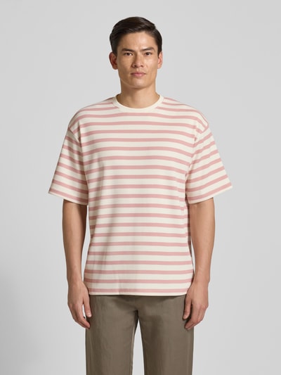 Männermodel trägt Oversized T-Shirt mit Rundhalsausschnitt Modell 'Gael' von Redefined Rebel, rosa-weiß gestreift, Elastischer-Material-Mix, Oversized-Fit, Rückenlänge bei Größe M 75 cm, Ganzkörperansicht von vorne.