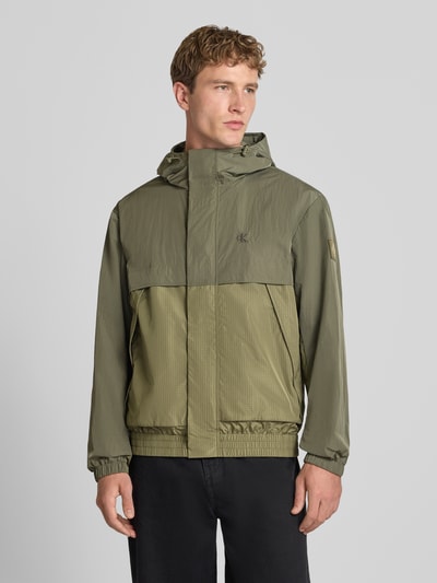 Männermodel trägt Regular Fit Regenjacke mit Rückenkoller von Calvin Klein Jeans w kratę, jednokolorowy, karczek, długi rękaw, wodoszczelny, wiatroszczelny, boczne kieszenie zapinane na zamek błyskawiczny, kieszeń wewnętrzna, kaptur z tunelem, wyhaftowane logo, naszywka z logo, elastyczne wykończenia, całkowity widok z przodu