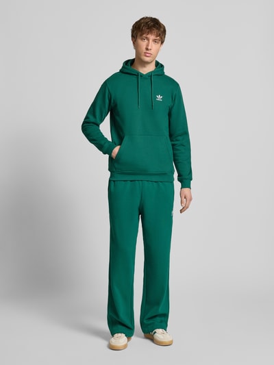 Männermodel trägt Wide Fit Sweathose mit elastischem Bund von adidas Originals in Grün, Baumwoll-Mix, Logo-Stitching, Eingrifftaschen, Ganzkörperansicht von vorne.