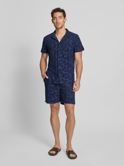 Mężczyzna model nosi Relaxed Fit Shorts aus Frottee mit Gesäßtasche od Scotch & Soda, mieszanka bawełny, krój relaxed fit, frota, elastyczny pas, wpuszczane kieszenie, kieszeń z tyłu, detal z logo, szerokość w rozmiarze M: 41 cm, długość wewnętrzna nogawki w rozmiarze M: 18 cm, granatowy, faktura froty, całkowity widok z przodu.