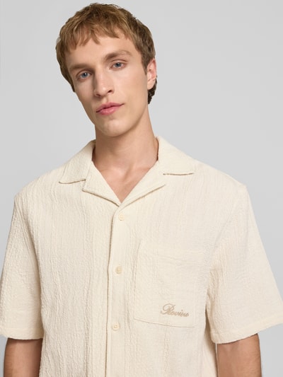 Męski model nosi Regular Fit Freizeithemd mit Logo-Stitching od REVIEW, kremowa bawełna, kieszeń, krótki rękaw, krój regular fit, zbliżenie na górną część ciała