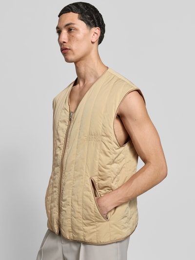 Männermodel trägt Relaxed Fit Weste in Stepp-Optik von MM6 Maison Margiela in Beige mit Eingrifftaschen, V-Ausschnitt, Reißverschluss, Ganzkörperansicht von vorne.