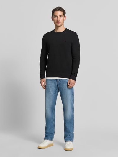 Männermodel trägt Regular Fit Pullover aus reiner Baumwolle von Tommy Hilfiger in Schwarz, kombiniert mit blauer Jeans, Ganzkörperansicht von vorne. Herren Pullover von Tommy Hilfiger.