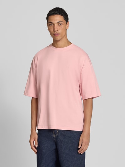 Męski model nosi Oversized T-Shirt aus reiner Baumwolle od REVIEW, różowy kolor, okrągły dekolt, nadruk z logo, bawełna, krój oversized, długość tyłu w rozmiarze M: 66 cm, widok całej sylwetki od przodu.