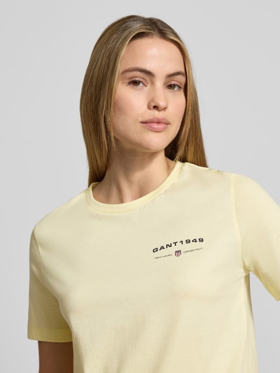 Modelka kobieca nosi T-Shirt z label-printem i okrągłym dekoltem marki Gant w kolorze jasnożółtym, czysta bawełna, regular fit, zbliżenie na górną część ciała.