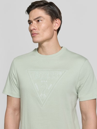 Männermodel trägt T-Shirt mit Logo und Rundhalsausschnitt von Guess aus Reine Baumwolle, Regular-Fit, dezente grüne Farbe, Nahaufnahme Oberkörper. Rückenlänge bei Größe M: 70 cm.