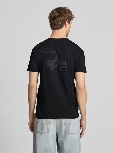 Männermodel trägt T-Shirt mit Label-Patch von Alpha Industries, reine Baumwolle, prosty krój, czarny kolor, widok całej sylwetki od tyłu.