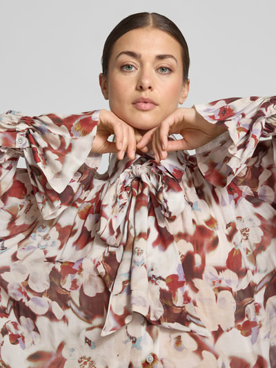 Frauenmodel trägt Bluse mit Schleife Modell 'VAELIE' von DRYKORN, florales Muster in Rot-Creme, lockerer Schnitt, Nahaufnahme Oberkörper. Material-Mix mit Modal-Anteil, Umlegekragen, Raglanärmel, elastische Bündchen, Größe 36: Rückenlänge 58 cm, Ärmellänge 62 cm.