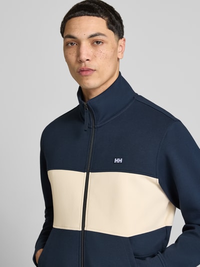 Męskimodel nosi bluzę rozpinaną z mieszanki bawełny z dodatkiem streczu model 'KOSTER' od Helly Hansen z naszywką z logo, prosty krój, stójka, zamek błyskawiczny, wpuszczane kieszenie, granatowo-beżowe bloki kolorystyczne, długość tyłu 67 cm, długość rękawów 66 cm, zdjęcie: z przodu, ujęcie do połowy torsu