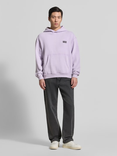 Męski model nosi fioletową bluzę z kapturem z logo, kieszenią kangurka z bawełny, krój relaxed fit, Hoodie mit Logo Modell 'PLIZZY' von American Vintage, długość tyłu 66 cm, długość rękawów 60 cm, Ganzkörperansicht von vorne.
