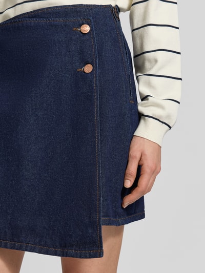 Frauenmodel trägt Regular Fit Minirock in Wickel-Optik Modell 'Lisana' von YAS, Denim-Optik, reine Baumwolle, 2-Knopf-Leiste, Nahaufnahme Oberkörper.
