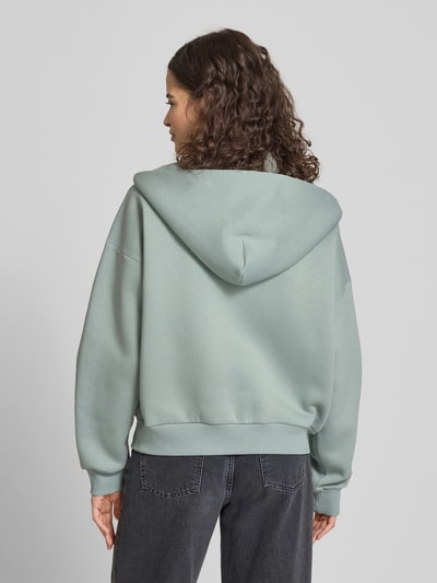 Frauenmodel trägt Sweatjacke mit Logo und Känguru-Tasche von Review in Mintgrün aus Baumwoll-Mix, Relaxed Fit, Ganzkörperansicht Rückansicht.