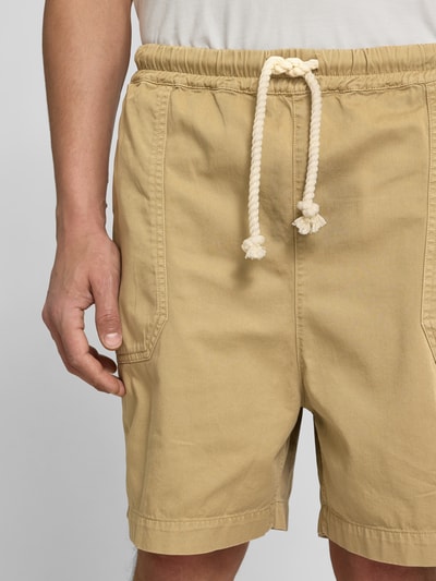 Männermodel trägt Bermudas aus reiner Baumwolle von Officine Générale in Beige mit elastischem Bund, Eingrifftaschen, Tunnelzug, Nahaufnahme Oberkörper.