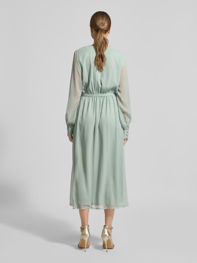 Frauenmodel trägt Midikleid mit Taillenband Modell 'FALIA' von VILA in semitransparenter Optik, weich fließender Stoff, Ganzkörperansicht Rückansicht. Taillierte Passform, Wickel-Optik, V-Ausschnitt, Blickdichter Unterrock, langärmelig, Rückenlänge 127 cm, Brustweite 47 cm, Ärmellänge 66 cm, pastellgrün.