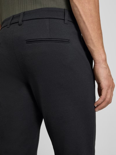 Mężczyznamodel nosi Slim Fit Hose mit Viskose-Anteil in verkürzter Passform von Lindbergh, czarne, gładka tekstura, wpuszczana kieszeń z tyłu, mieszanka wiskozy, krój slim fit, skrócona długość, szerokość w rozmiarze M: 42 cm, długość wewnętrzna nogawki: 72 cm, elastyczna mieszanka materiałów, zapięcie na guzik i zamek błyskawiczny, kieszenie wsuwane, widok z tyłu, detal.