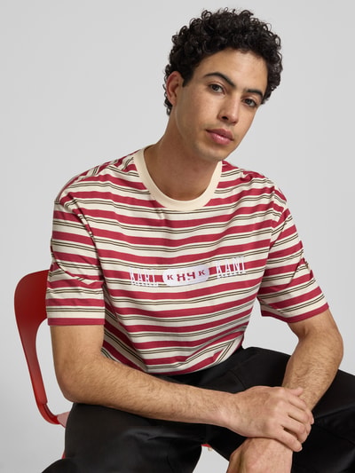 Męski model nosi Relaxed Fit T-Shirt mit Logo-Stitching Modell 'Heritage' od KARL KANI, bawełna, krój relaxed fit, okrągły dekolt, długość tyłu w rozmiarze M 64 cm, czerwone i beżowe paski, miękka tekstura, nahaufnahme oberkörper.