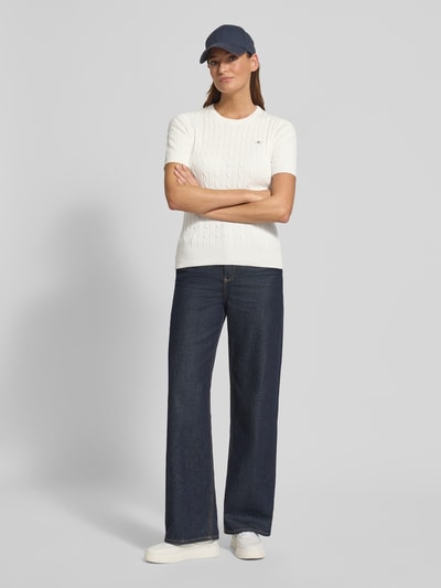 Kobiece modelka nosi Strickshirt mit Zopfmuster und Label-Stitching od Gant, mieszanka bawełny, krój slim fit, okrągły dekolt, haftowane logo, kolor biały, ciemnoniebieskie spodnie, długość tyłu 57 cm, długość rękawów 24 cm, całkowity widok z przodu.