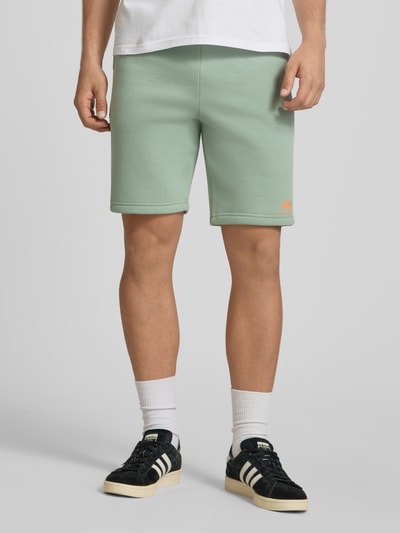 Männermodel trägt Shorts mit Eingrifftaschen und Logo-Print von Alpha Industries in Mintgrün, Relaxed Fit, Ganzkörperansicht von vorne.