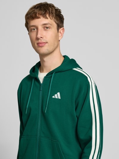 Männermodel trägt Regular Fit Sweatjacke aus Baumwoll-Mix von adidas Sportswear in Dunkelgrün mit weißen Streifen, Nahaufnahme Oberkörper.