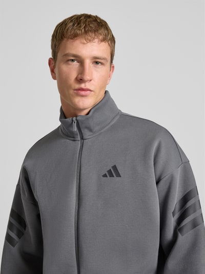 Männermodel trägt Sweatjacke mit Logo und Reißverschluss von adidas Sportswear, grauer Material-Mix, Stehkragen, Nahaufnahme Oberkörper.