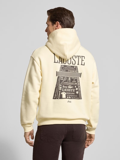 Männermodel trägt Regular Fit Hoodie aus reiner Baumwolle von Lacoste w kolorze jednolitym z nadrukiem z motywem oraz naszywką z logo, krój regular fit, kaptur, długie rękawy z prążkowanym wykończeniem, kieszeń kangurka, dzianina dresowa, materiał: bawełna, widok całej sylwetki z tyłu.