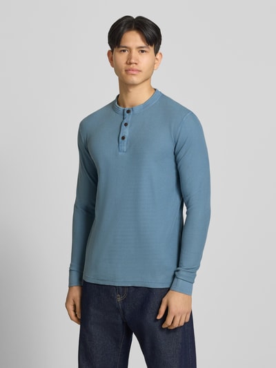 Męski model nosi Longsleeeve mit Knopfleiste od Superdry, niebieska bawełna, krój regular fit, okrągły dekolt, długość tyłu 72 cm, Ganzkörperansicht von vorne.