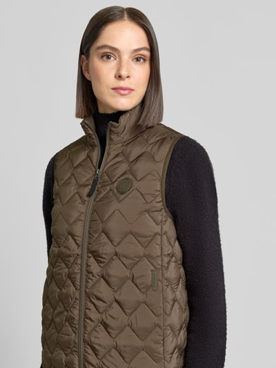 Kobiece modelka nosi kamizelkę Regular Fit w kolorze oliwkowym z efektem pikowania model 'Evy' od Didriksons, detal z logo, zamek błyskawiczny, stójka, Długość tyłu w rozmiarze 36: 63 cm, Szerokość ramion w rozmiarze 36: 37 cm, Szerokość klatki piersiowej w rozmiarze 36: 46 cm, półzbliżenie przód
