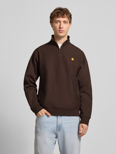 Männermodel trägt Sweatshirt mit Label-Stitching von Carhartt Work In Progress in Braun aus Baumwoll-Mix, lockerer Schnitt, Stehkragen, kurzer Reißverschluss, gerippte Abschlüsse, Rückenlänge 70 cm, Ärmellänge 64 cm, Ganzkörperansicht von vorne.