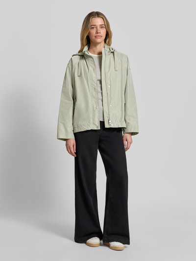 Frauenmodel trägt Jacke mit Kapuze Modell 'CICATA' von CINQUE in hellem Grün, glatte Textur, seitliche Taschen, Ganzkörperansicht von vorne.