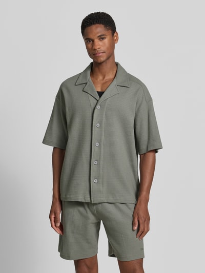 Męski model nosi Relaxed Fit Freizeithemd mit Strukturmuster von Drykorn z bawełny, kołnierz z połami, krótki rękaw, listwa guzikowa, szerokość ramion w rozmiarze M 60 cm, długość tyłu 69 cm, długość rękawów 26 cm, fakturowany wzór, widok całej sylwetki z przodu.