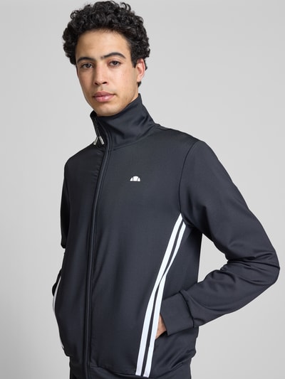 Männermodel trägt Sweatjacke mit Reißverschlusstaschen Modell 'TOSCIANA' von Ellesse, Schwarz, Logo-Print, Ganzkörperansicht von vorne. Material-Mix mit Stretch-Anteil, Gerader-Schnitt, Stehkragen, Reißverschluss, seitliche Reißverschlusstaschen, elastische Abschlüsse, Rückenlänge 69 cm, Ärmellänge 70 cm.