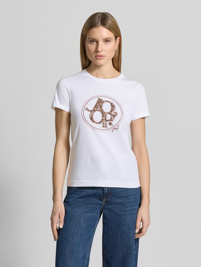 Kobiecy model nosi damski t-shirt z bawełny o prostym kroju, okrągłym dekolcie, obszyciu z kamieni stras i detalem z logo; t-shirt z kolekcji JOOP!, długość tyłu w rozmiarze 36: 59 cm, widok całej sylwetki z przodu.