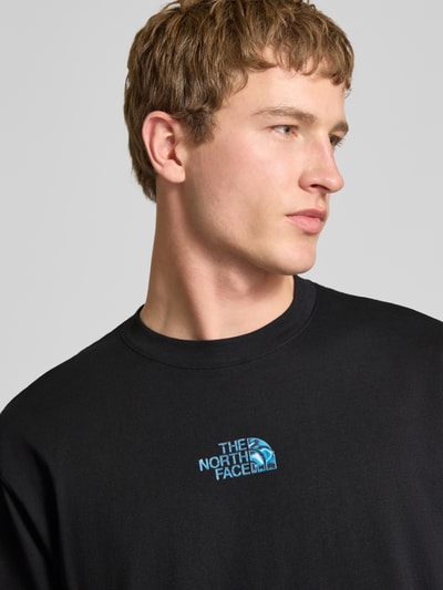 Mężczyznę-model nosi Oversized T-Shirt mit Logo und Rundhalsausschnitt von The North Face, czarna bawełna, logo z przodu, oversize, długość tyłu w rozmiarze M: 73 cm, zbliżenie na górną część ciała