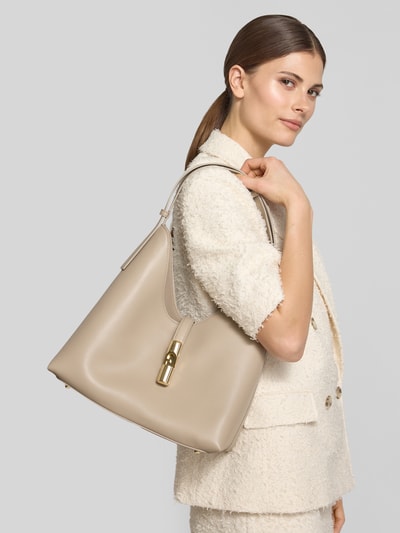 Frauenmodel trägt Handtasche mit Logo-Detail Modell 'Goccia' von FURLA in Beige, Oberflächenstruktur glatt, Ganzkörperansicht von vorne, Hauptfach mit Riemenverschluss, Verstellbarer Trageriemen, Breite 25 cm, Länge 31 cm, Tiefe 15 cm, Schulterriemenlänge 42 cm.