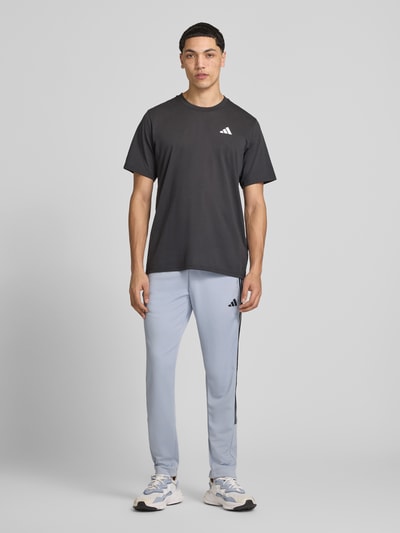 Männermodel trägt Slim Fit Sweatpants mit Logo-Print von adidas Training, hellgrau, mit Reißverschluss-Eingrifftaschen und elastischem Bund, Ganzkörperansicht von vorne.