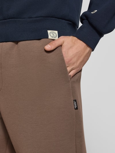 Männermodel trägt Regular Fit Sweatpants aus Material-Mix Modell 'BILL LEGACY' von JACK & JONES in Braun, Nahaufnahme Hüfte, Herren Sweatpants mit Eingrifftaschen und elastischem Bund mit Tunnelzug, Innenbeinlänge bei Größe M 77 cm.