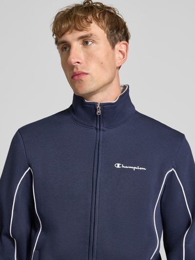 Männermodel trägt Trainingsanzug aus Baumwoll-Mix von Champion in Dunkelblau mit weißen Kontrastnähten und Label-Print, Nahaufnahme Oberkörper.