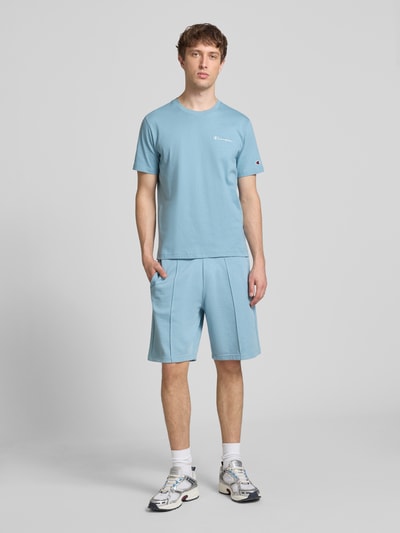 Męski model trägt Bermudas mit elastischem Bund und Label-Stitching von CHAMPION, mieszanka bawełny, krój regular fit, jasnoniebieski, szwy podłużne, wyhaftowane logo, szerokość w rozmiarze M 42 cm, długość wewnętrzna nogawki w rozmiarze M 24 cm, ganzkörperansicht von vorne.