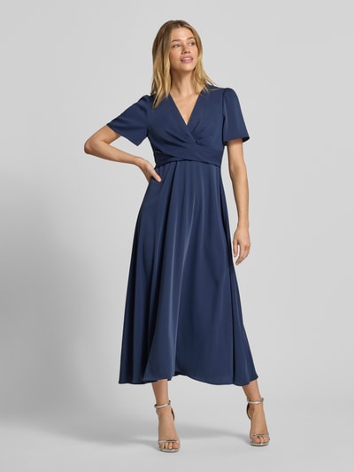 Frauenmodel trägt Cocktailkleid mit V-Ausschnitt von Christian Berg Cocktail in Dunkelblau, A-Linie, Midilänge, Knotendetail, Ganzkörperansicht von vorne.