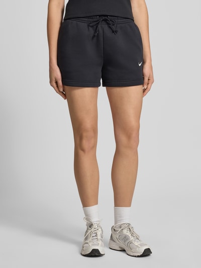 Frauenmodel trägt Sweatshorts mit Logo Modell 'Phoenix' von Nike, Baumwoll-Mix, schwarzer Stoff, Logo links, Ganzkörperansicht von vorne.
