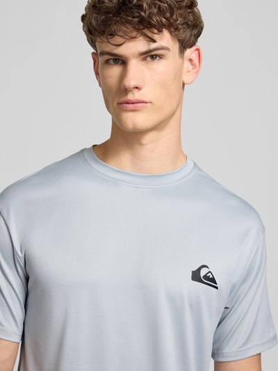Männermodel trägt T-Shirt mit Rundhalsausschnitt und Logo von Quiksilver, hellgrau, Regular Fit, Material-Mix mit Stretch, Nahaufnahme Oberkörper.