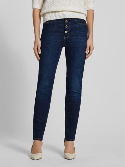 Kobieta nosi Jeansy z 5 kieszeniami od Guess, mieszanka bawełny ze streczem, krój slim fit, zapięcie na guzik, ciemnoniebieskie; całkowity widok z przodu.