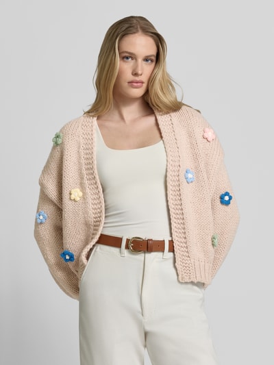 Frauenmodel trägt Oversized Strickjacke mit floralem Zierbesatz von Apricot, Grobstrick, offene Vorderseite, Gerippte-Abschlüsse, Ganzkörperansicht von vorne. Rückenlänge 60 cm, Ärmellänge 51 cm, überschnittene Schultern, Damen.