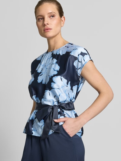 Modelka nosi Blusenshirt aus Satin mit Bindegürtel von Christian Berg Cocktail w granatowym kolorze z jasnoniebieskim kwiatowym wzorem, krój taliowany, tkanina satynowa, pasek w talii, okrągły dekolt, tekstura gładka, cały look, perspektywa: zbliżenie na górną część ciała.