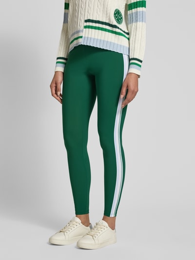 Kobiece model nosi Slim Fit Leggins mit elastischem Bund od Marc Cain, elastyczna mieszanka materiałów, krój slim fit, ozdobne obszycie, szerokość w rozmiarze 36: 34 cm, długość wewnętrzna nogawki w rozmiarze 36: 76 cm, zielony kolor z białym paskiem, zdjęcie całej sylwetki z przodu.