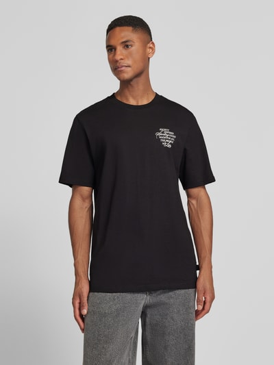 Męsk model nosi T-Shirt mit rückseitigem Motiv-Print Modell 'Ben Faience' od Les Deux, czarny kolor, bawełna, prosty krój, widok z przodu na całą sylwetkę.