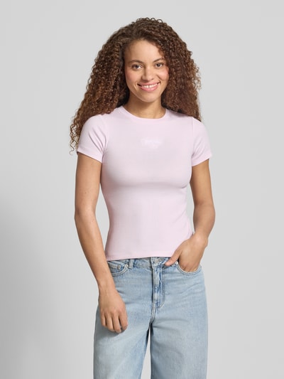 Frauenmodel trägt T-Shirt in Ripp-Optik mit Logo von Guess Jeans in zartem Rosa, Slim Fit, Rundhalsausschnitt, Ganzkörperansicht von vorne.